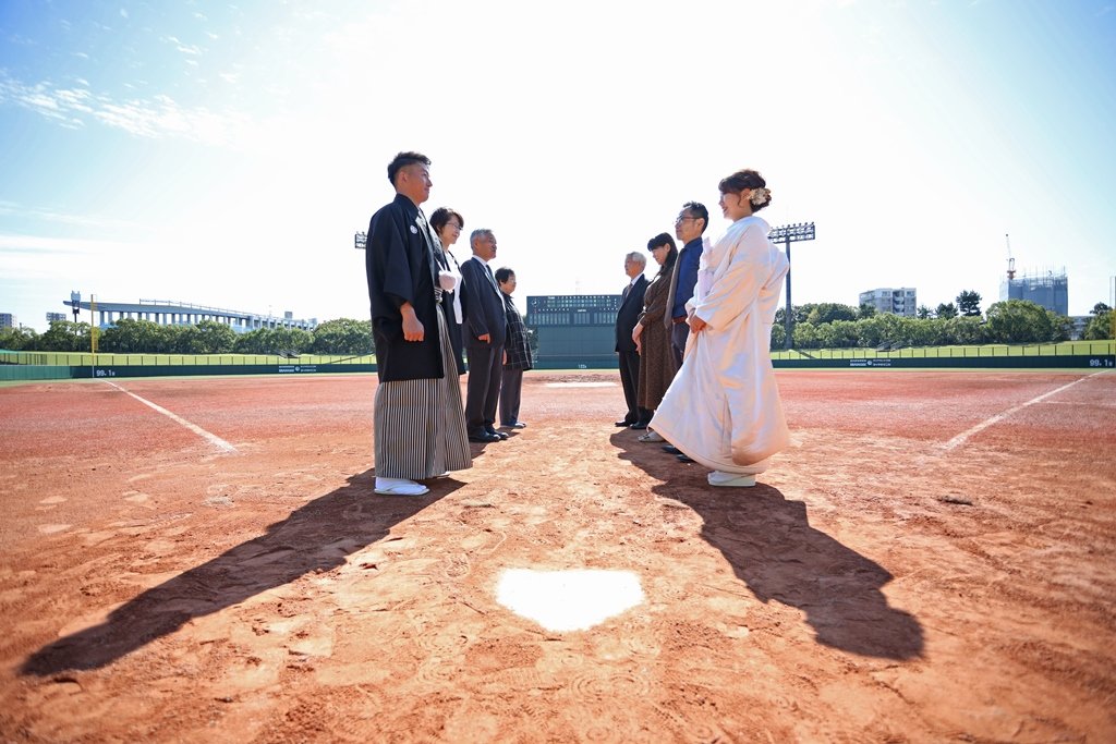 野球場のホームベースを挟んで向かい合う黒紋付袴の新郎と白無垢姿の花嫁、両脇には見守る家族の姿