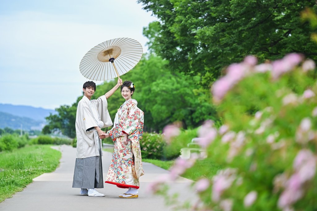 ピンク色の花が咲く鴨川沿いの遊歩道で、白い和傘を高くかかげ手をつなぐ生成り色の着物袴の新郎と花柄着物の花嫁