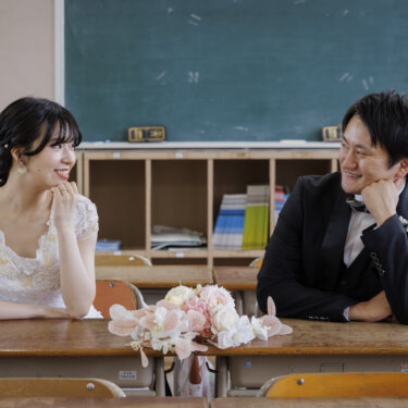 教室の机に座って頬杖をつき、見つめ合うレースのウェディングドレスの花嫁と黒いタキシードの新郎。母校の教室で撮る洋装前撮り