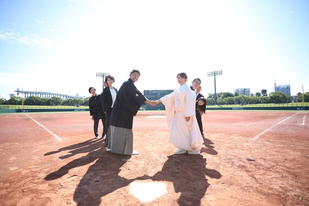 野球場のホームベース上で握手を交わす黒紋付袴の新郎と白無垢姿の花嫁、後ろから顔をのぞかせる家族たち