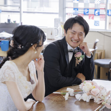 教室の窓辺の机で笑い合うレースのウェディングドレスの花嫁と黒いタキシードの新郎。学校ロケーションで撮る洋装前撮りの教室カット