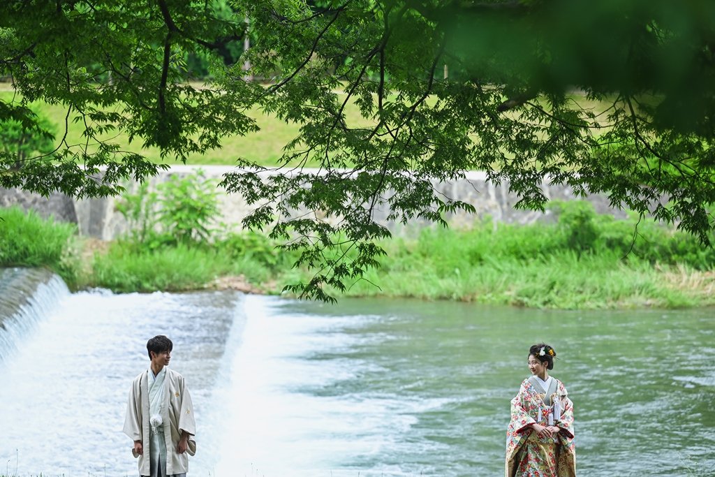 鴨川の堰から水が流れる中、川の中に立って向かい合う生成り色の着物袴の新郎と花柄着物の花嫁