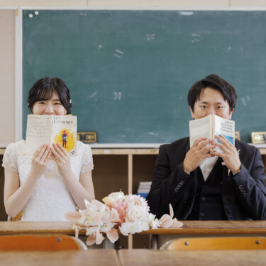 黒板を背にした机で、文庫本で顔を半分隠してこちらを見る新郎新婦。教室でのユニークなウェディングフォト