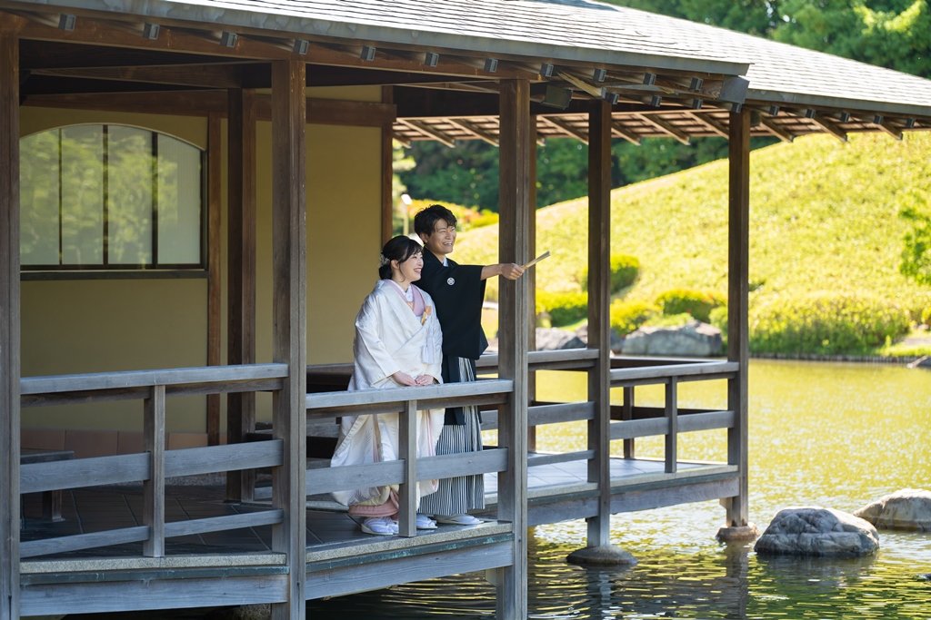 池に面した木造の四阿で扇子を掲げる黒紋付袴の新郎と白無垢の花嫁