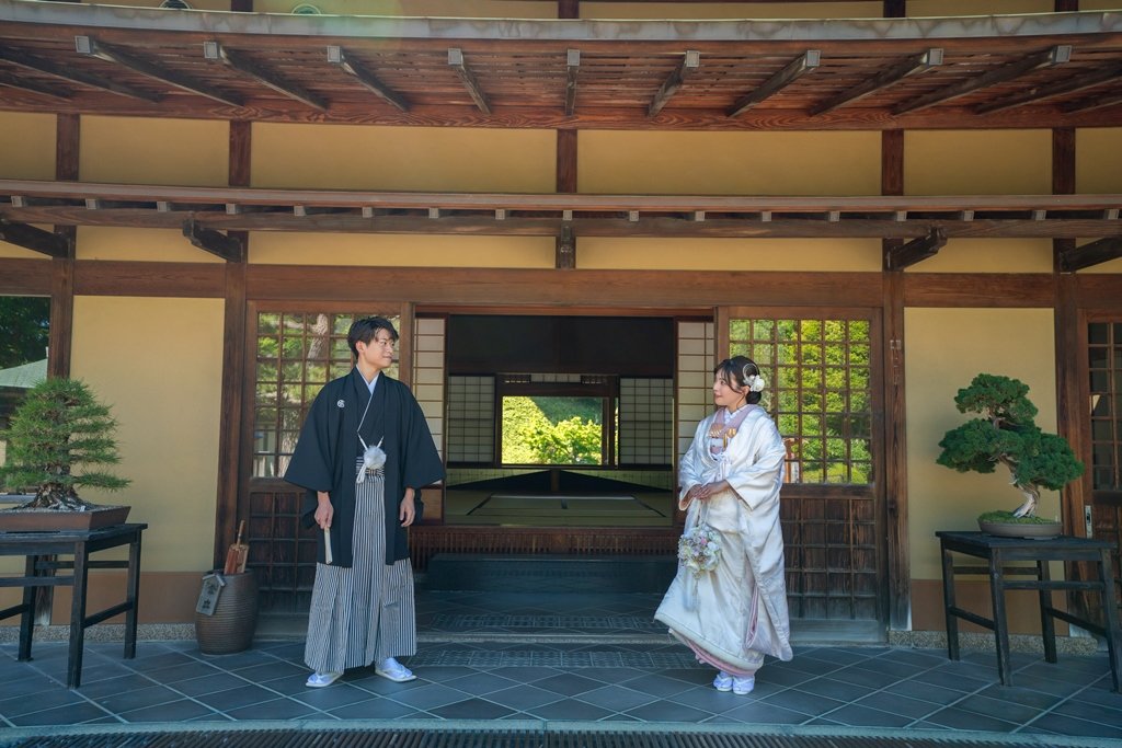 盆栽が飾られた玄関先で見つめ合う、黒紋付袴の新郎と白無垢にブーケを持った花嫁