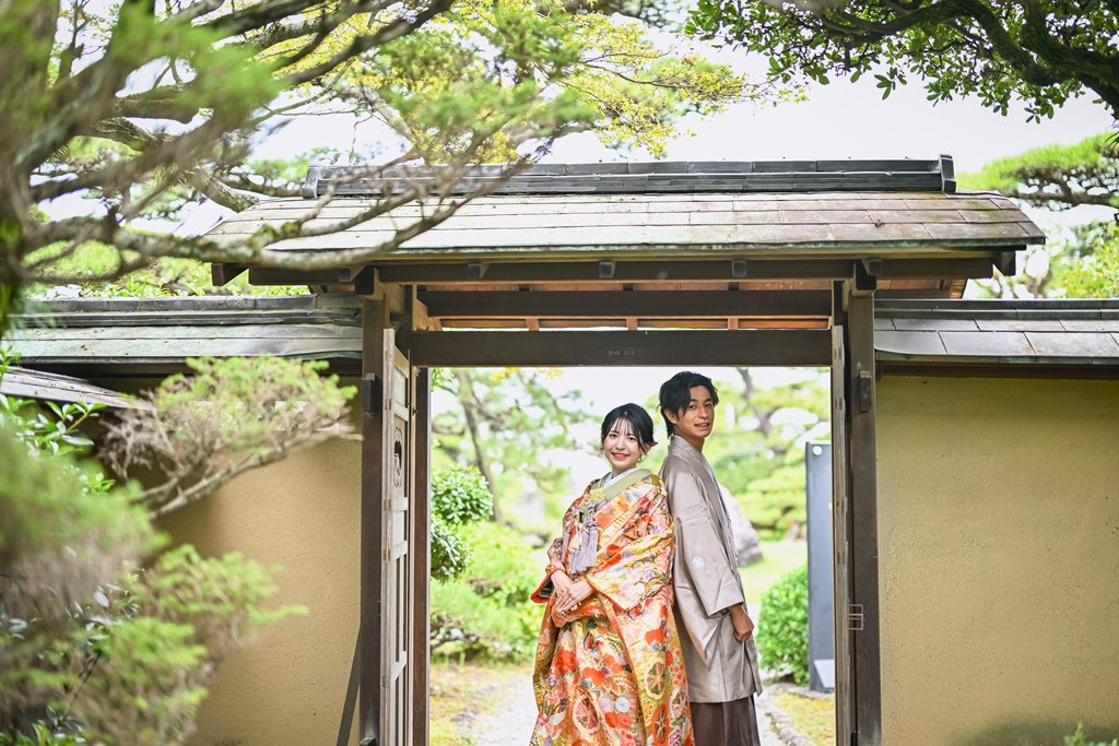 瓦屋根の木造門の下、木々に囲まれ背中合わせに立つ朱色の花柄着物の花嫁と灰色の着物姿の新郎