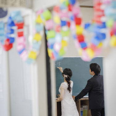 色とりどりの輪飾りを前ボケにして、黒板に文字を書くウェディングドレスの花嫁とタキシードの新郎をとらえた一枚。母校の教室で撮る洋装前撮り