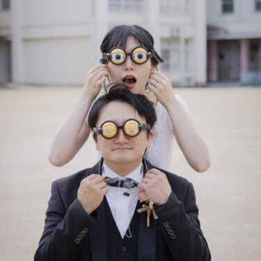 校庭で、おもしろい眼鏡をかけたウェディングドレスの花嫁とタキシードの新郎が重なって笑う縦位置の一枚。学校ロケーションで撮る遊び心のある前撮り