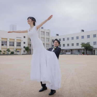 校庭で新婦を横抱きにして持ち上げ、両手を広げて笑う新郎新婦。校舎を背にした学校ロケーションフォト