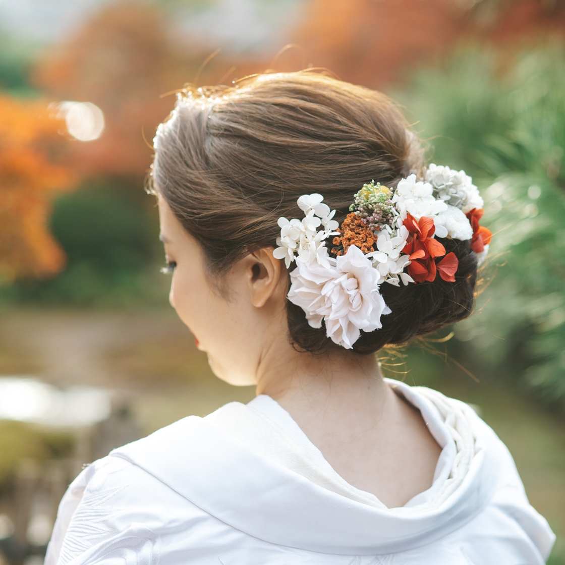紅葉を背景に赤と白の生花を華やかに飾った、和装向けアップスタイルのヘアメイク