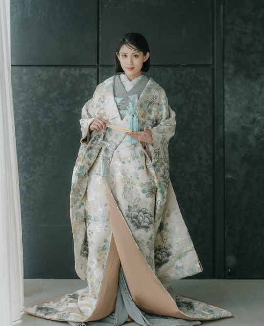 ベージュの生地にニュアンスブルーやゴールドが調和し、気品あるクールさを演出する色打掛。繊細な彩りがモダンに重なり合い、奥行きを感じさせる洗練のデザイン。裏地には珍しいブルーをあしらい、トレンド感と個性をさりげなく添える。静かな輝きが花嫁の美しさを際立たせる上質な一着。（柔韻麗景／衣装番号AMOS18）