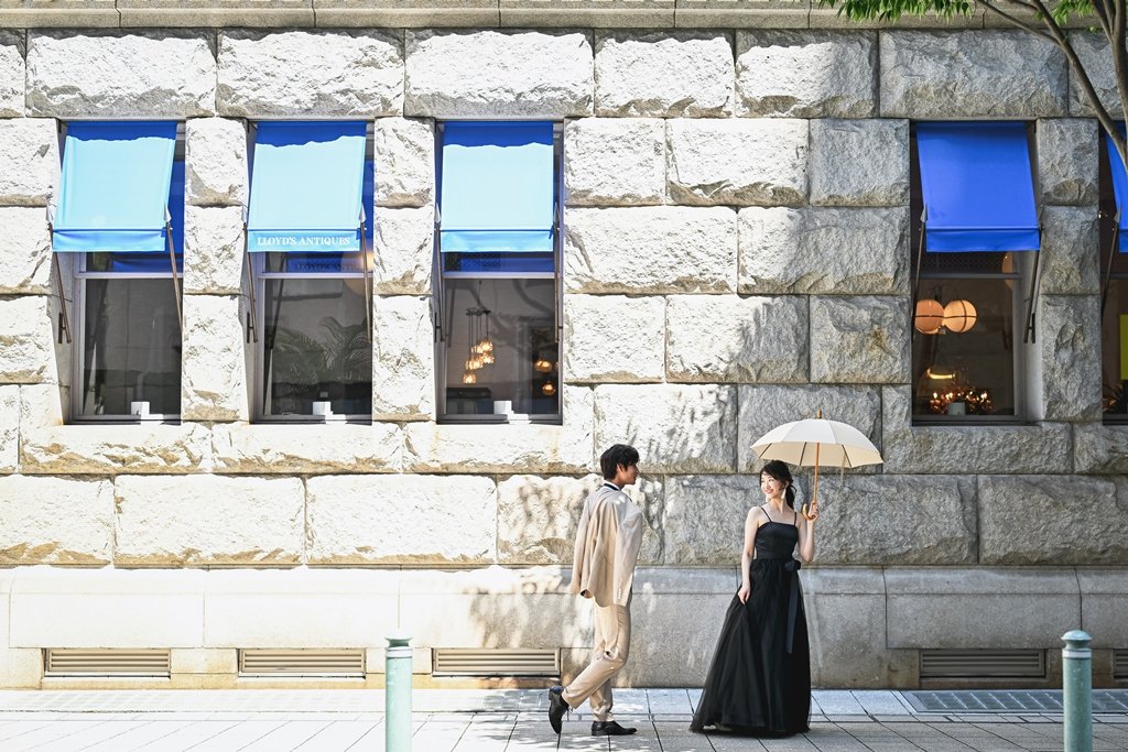 青いオーニングが並ぶ神戸旧居留地の石造りの建物の前を歩く、ベージュのスーツの新郎と、白いパラソルをさす黒いドレスの花嫁