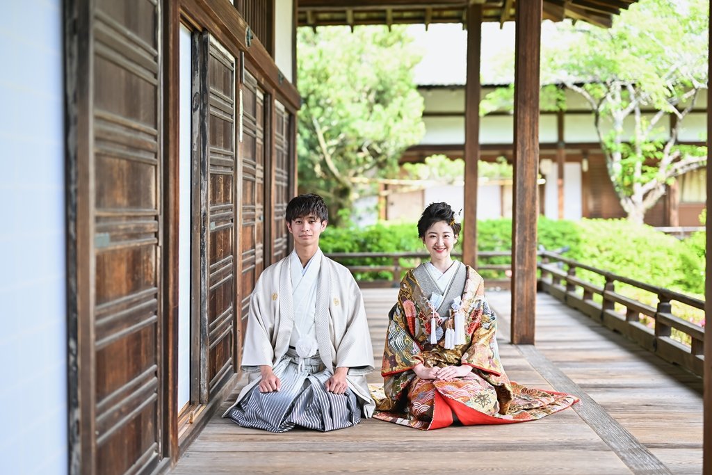毘沙門堂の木造の渡り廊下で正座して並ぶ、生成り色の着物袴の新郎と、朱色の裏地が美しい豪華な花柄着物の花嫁