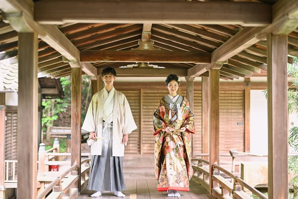 木造の梁が美しい毘沙門堂の渡り廊下を歩く、生成り色の着物袴の新郎と朱色の裏地が覗く花柄着物の花嫁