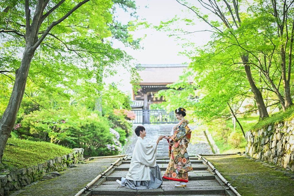 新緑のもみじが両側に茂る参道の石段で、片膝をつき花嫁の手を取る生成り色の着物姿の新郎と豪華な花柄着物の花嫁