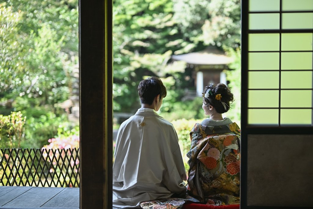 障子越しに新緑の庭園を望む縁側で、座布団に正座して庭を眺める後ろ姿の新郎新婦
