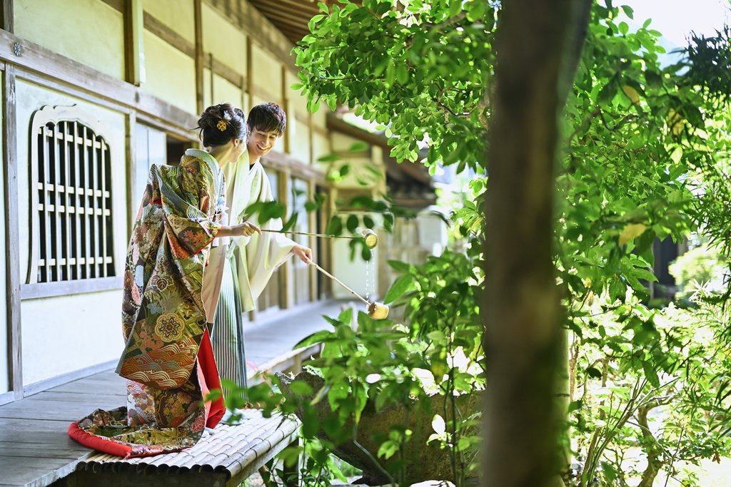 緑豊かな毘沙門堂の縁側で、柄杓を手にした生成り色の着物袴の新郎と朱色の花柄着物を着た花嫁が手水を使う