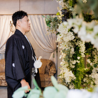 装花越しに横向きで立つ黒紋付袴の新郎。洋風のソファとランプを背景にした和装スタジオフォトの新郎カット