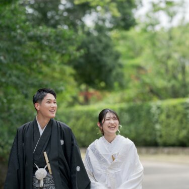 緑の生垣の小道を笑顔で並んで歩く紋付袴の新郎と白無垢の花嫁。夏の大仙公園で撮る自然体の和装フォトウェディング