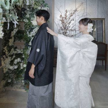 白い花のアーチの下で、新郎の肩にそっと手を伸ばす白無垢の花嫁。心斎橋店のスタジオで撮影した和装フォトウェディング