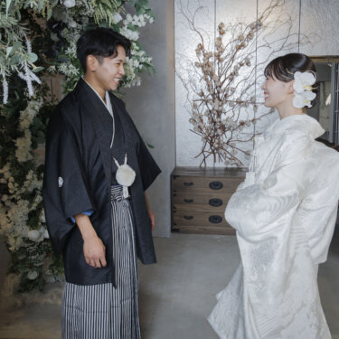 グリーンと白い花に囲まれたスタジオで、向かい合って笑い合う紋付袴の新郎と白無垢の花嫁の前撮り