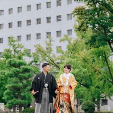芝生の広場を手をつないで歩く、紋付袴の新郎と金色の色打掛の花嫁。緑の木々に囲まれた和装ロケーションフォト
