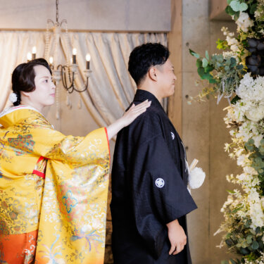 新郎の肩に手を添えて衿元を整える色打掛の花嫁。支度のひとこまのような自然な和装前撮り