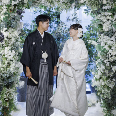 白い花とグリーンのフラワーアーチの下に並んで立ち、見つめ合う新郎新婦。大阪・心斎橋の和装スタジオフォト