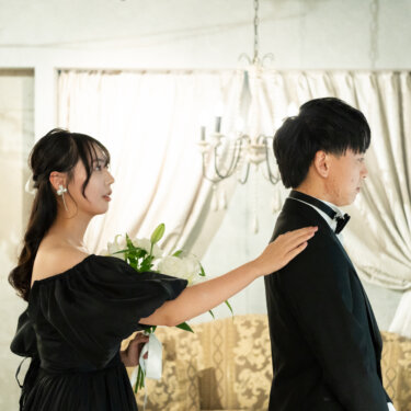 後ろを向いた黒いタキシードの新郎の肩に、ユリのブーケを持った黒のカラードレスの花嫁がそっと手を置くファーストミートのポーズ。大阪のスタジオでの前撮り