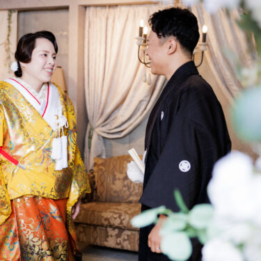カーテンとソファのある洋風セットで顔を見合わせて笑う色打掛の花嫁と紋付袴の新郎。和洋ミックスの前撮り