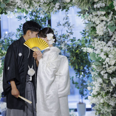 フラワーアーチの前で、新郎が金色の扇子で二人の顔を隠すキスシーン風の和装前撮りポーズ