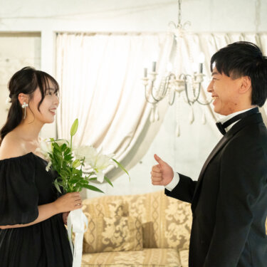 シャンデリアのある部屋で、ブーケを持つ黒のカラードレスの花嫁に親指を立てて笑う新郎。ファーストミートの瞬間を撮った前撮りのワンシーン