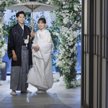 花のアーチの下で白い和傘を差して並ぶ紋付袴の新郎と白無垢の花嫁。大阪の心斎橋店で撮影した結婚写真