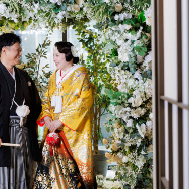 窓越しにのぞくように撮った、フラワーアーチの下で向き合う新郎新婦。自然な会話が伝わる和装スタジオフォト