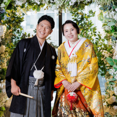 白い花のフラワーアーチの下で並んでほほえむ紋付袴の新郎と色打掛の花嫁。大阪のスタジオで撮る和装前撮り