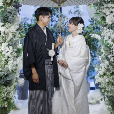 白い和傘の下で見つめ合う新郎新婦を、フラワーアーチ越しに縦構図で撮った和装スタジオフォト