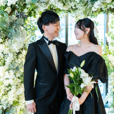 グリーンと白い花のフラワーアーチの前で、ユリのブーケを持って見つめ合う黒いタキシードの新郎と黒のカラードレスの花嫁。心斎橋店のスタジオフォト
