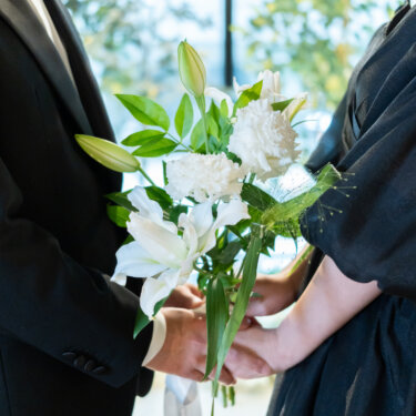 ユリと白い花のブーケを一緒に持って手を重ねる、黒いタキシードの新郎と黒のカラードレスの花嫁の手元。窓からの光がやさしい前撮りのカット