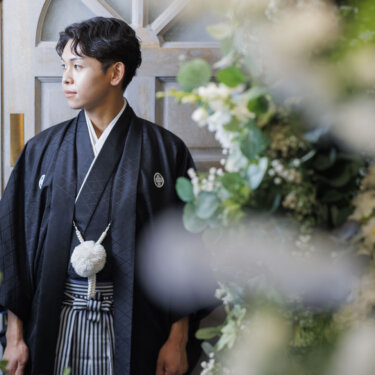 花やグリーンを手前にぼかし、遠くを見つめる黒紋付羽織袴の新郎のソロカット。和装前撮りの一枚