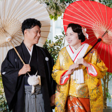白と赤の和傘を手に、グリーンの装花の前で見つめ合う紋付袴の新郎と黄色の色打掛の花嫁。和装前撮りの定番アイテム
