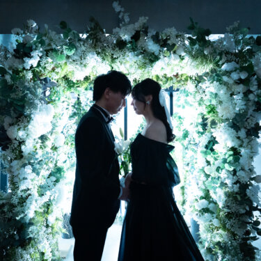 光る白い花のフラワーアーチの下で、おでこを寄せ合う新郎と黒のカラードレスの花嫁のシルエット。逆光を生かしたドラマチックな前撮り