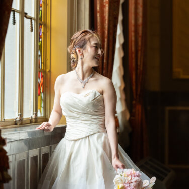 赤いカーテンとステンドグラスの窓辺で、ブーケを手に振り返るビスチェのウェディングドレスの花嫁の洋装前撮り