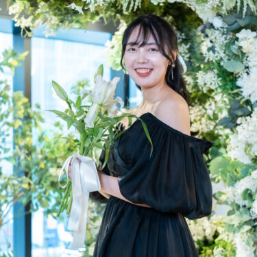 白とグリーンのフラワーアーチの前で、ユリのブーケを抱えて笑顔を見せるパフスリーブの黒いカラードレスの花嫁。リボンの髪飾りがかわいいスタジオフォト