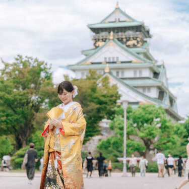 大阪城天守閣の前の広場で、色打掛の裾を広げてうつむき微笑む花嫁のソロカット。大阪城公園で撮る和装前撮り