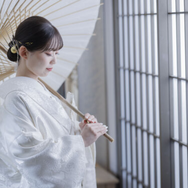 白い和傘を肩にかけ、格子窓の光の中で目を伏せる白無垢の花嫁。大阪の和装スタジオでの前撮り