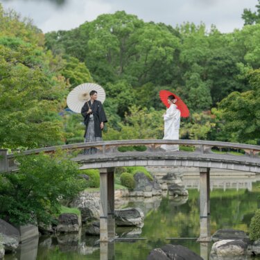 池にかかる反り橋の上で、赤と白の和傘を手に離れて立つ新郎新婦。大仙公園の日本庭園を広く写した和装前撮り