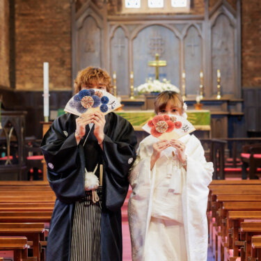 扇子で顔を半分隠して並ぶ紋付袴の新郎と白無垢の花嫁。レンガの壁と祭壇が背景の、教会で撮る和装前撮りの顔隠しポーズ