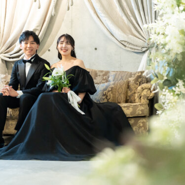 白い花のフラワーアーチ越しにソファの新郎新婦を写した、黒×黒コーディネートのスタジオフォト。黒のカラードレスの裾が床に広がる