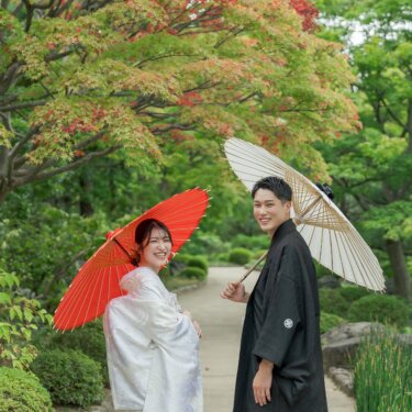 色づき始めたモミジの下で、赤い和傘の花嫁と白い番傘の新郎が振り返る。大仙公園の小道で撮る和装前撮りの人気ポーズ