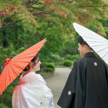 赤い和傘をさす白無垢の花嫁と、白い和傘を肩にかけて笑う新郎。モミジの葉が映える大仙公園の和装フォトウェディング